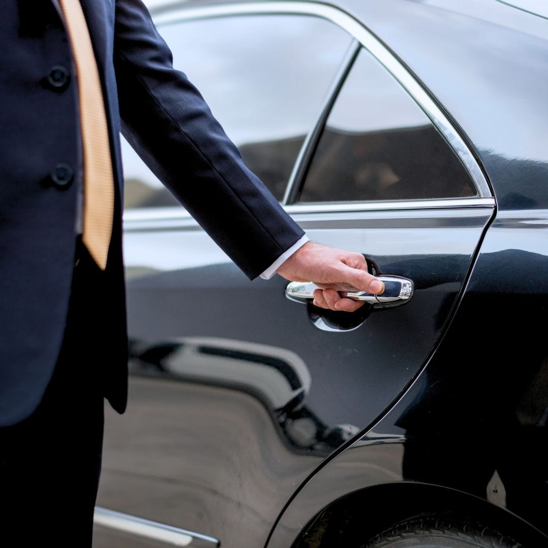 Chauffeur in a dark suit opening the door of a polished black luxury sedan for airport transfer.